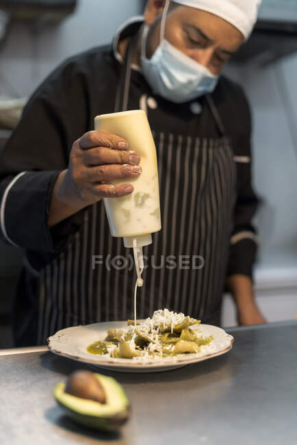 Crop middle aged ethnic male cook pouring white sauce from bottle on delicious pasta with grated cheese in restaurant kitchen — Stock Photo