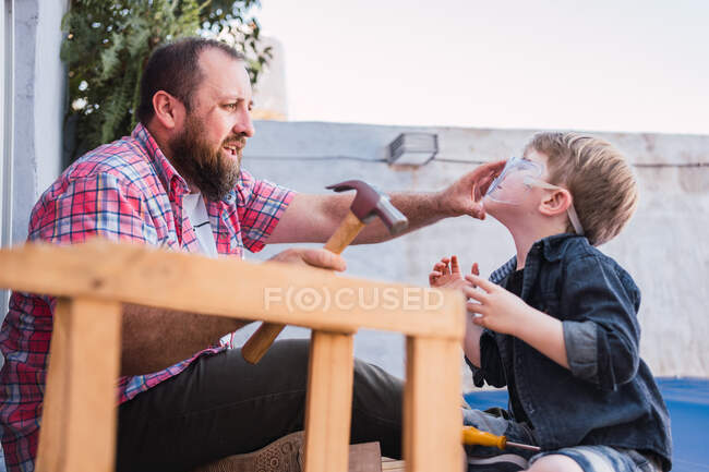Side view of hipster dad with hammer putting protective glasses on boy while preparing for woodwork — Stock Photo