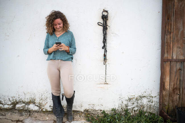 Interested female in riding boots text messaging on cellphone against stall with bridle on wall in daytime — Stock Photo