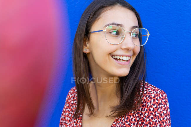 Glückliches Weibchen in stylischem roten Kleid steht tagsüber vor blauer Wand und schaut weg — Stockfoto