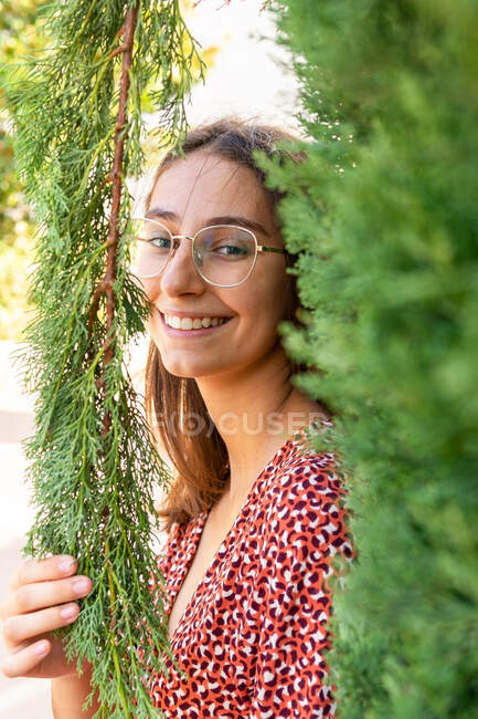 Fröhliche junge Frau mit braunen Haaren in Brille, die zwischen grünen Zweigen steht und bei Tageslicht in die Kamera schaut — Stockfoto