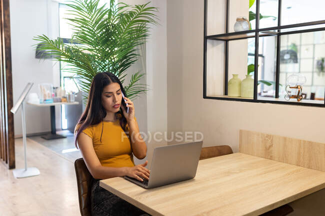 Serious female freelancer sitting at wooden table in cafe and having phone call while typing on netbook — Stock Photo