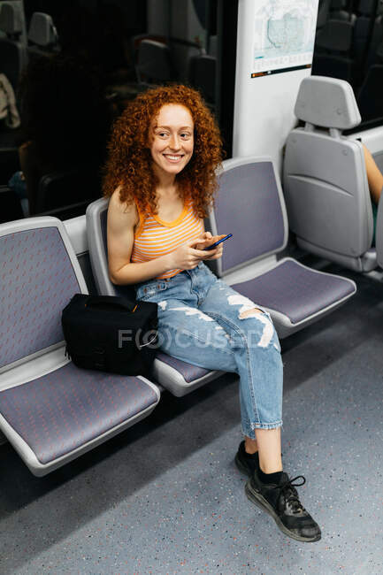 Femme intéressée avec les cheveux bouclés en jeans déchiré messagerie texte sur téléphone portable pendant le voyage en train en journée — Photo de stock