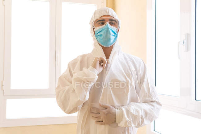 Médico adulto masculino em equipamento de proteção individual com óculos e máscara estéril olhando para a frente contra a janela no hospital — Fotografia de Stock