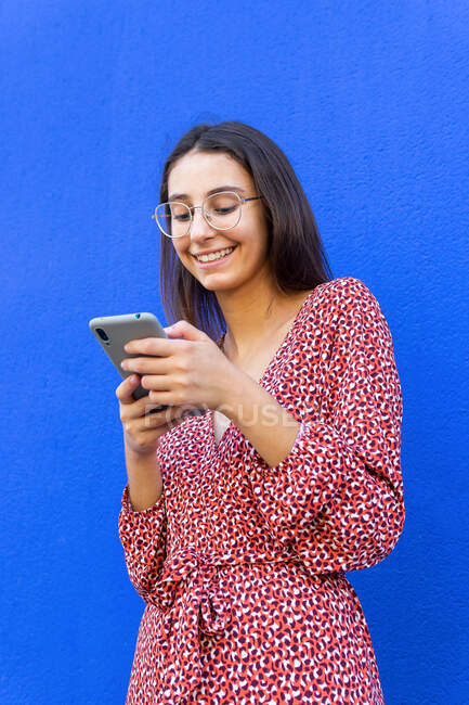 Smiling female in dress and eyeglasses standing near blue wall and using smartphone in daytime — Stock Photo