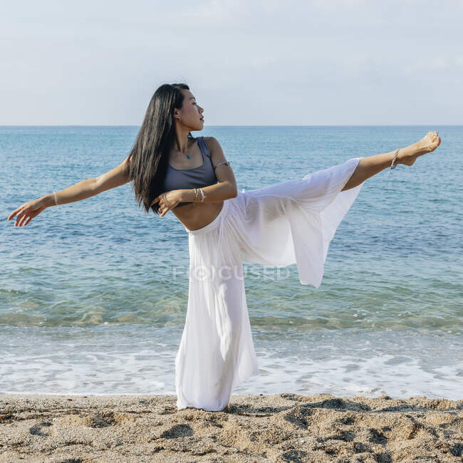 Hembra étnica con cabello largo balanceándose en una pierna mientras mira hacia otro lado en la orilla arenosa durante la práctica de yoga contra el mar - foto de stock