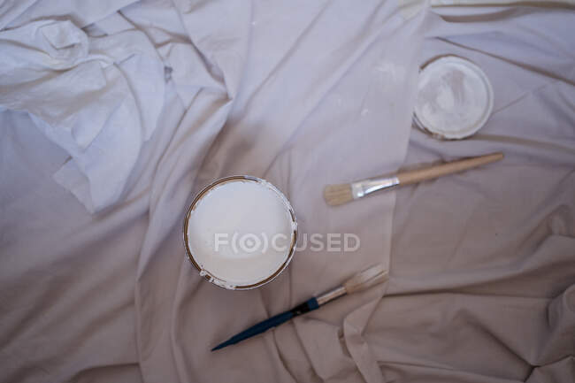 Vista superior del contenedor de pintura blanca con tapa y pinceles sobre tela arrugada durante la renovación en casa - foto de stock