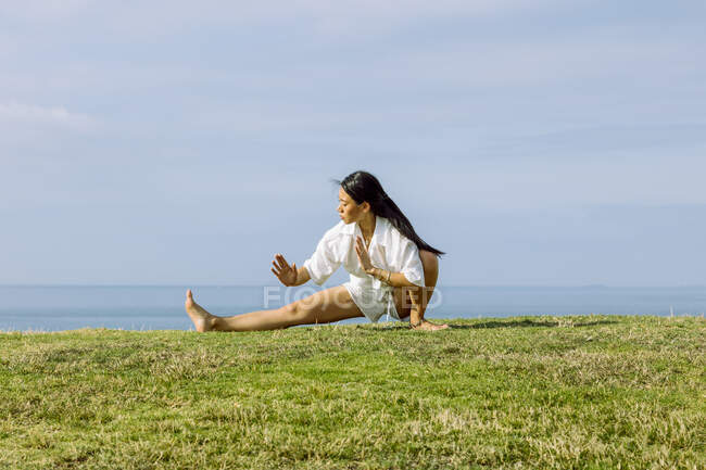 Молода етнічна самиця, яка сидить у позі Скандасани, практикуючи йогу на узбережжі трави проти моря під хмарним небом. — стокове фото