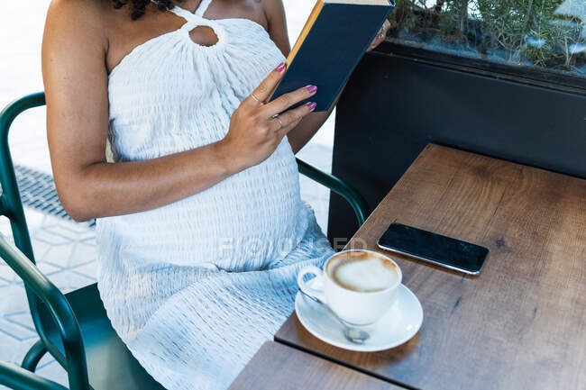 Cropped unrecognizable pregnant African American female with curly hair sitting at table and reading book while drinking coffee — Stock Photo
