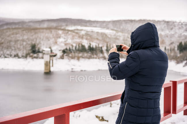 Back view of unrecognizable person standing near fence and taking photo of frozen river and snowy winter hills — Stock Photo
