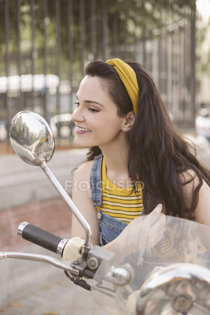 Encantadora jovem morena sentada na motocicleta e olhando no espelho retrovisor com sorriso de dente na rua — Fotografia de Stock