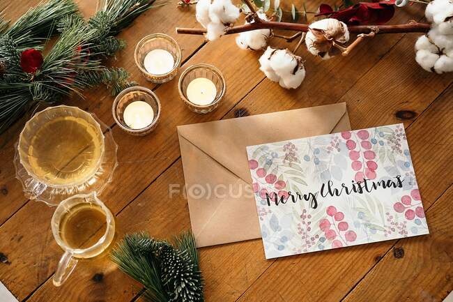 Vista superior de la composición navideña con postal colorida con inscripción Feliz Navidad colocada cerca de velas encendidas y tazas de té sobre una mesa de madera decorada con ramas coloridas de plantas - foto de stock