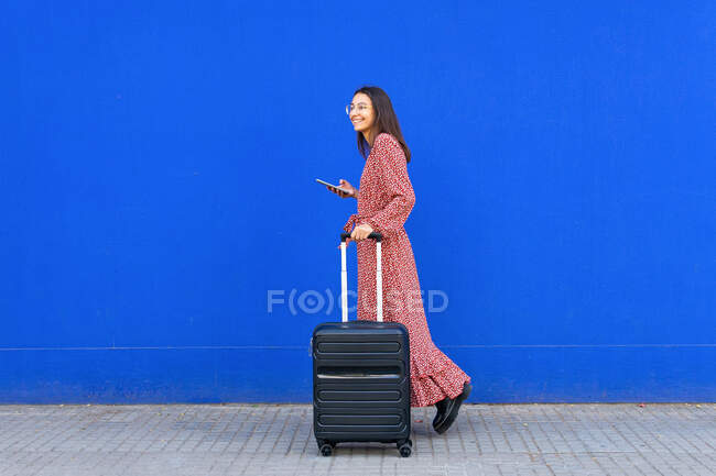 Vista laterale della femmina positiva in abito rosso lungo che cammina con i bagagli durante la navigazione sullo smartphone per strada contro il muro blu durante il giorno — Foto stock