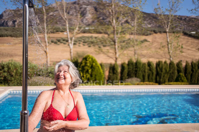 Веселая пожилая женщина в бикини наслаждается брызгами из душа у бассейна с прозрачной прозрачной водой — стоковое фото
