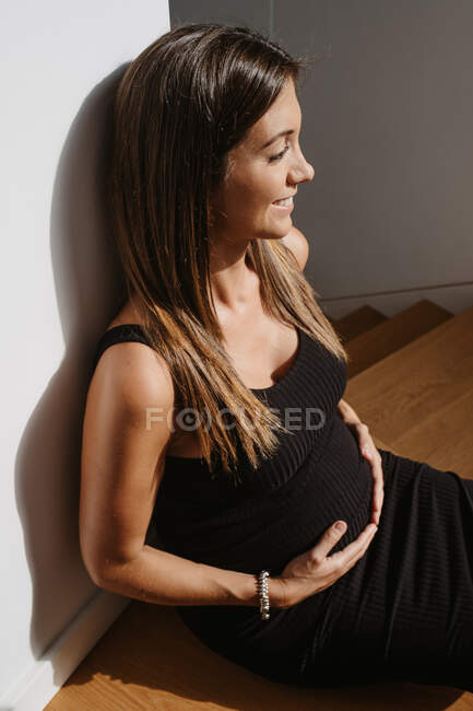 Dreamy adult expectant female stroking tummy while sitting on floor in house on sunny day looking away — Stock Photo