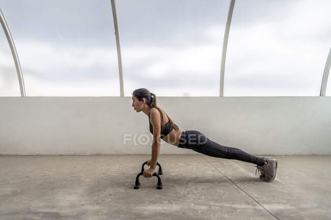 Вид сбоку на сидящую этническую женщину в активной одежде, выполняющую упражнения на отжимании штанги, глядя вперед — стоковое фото