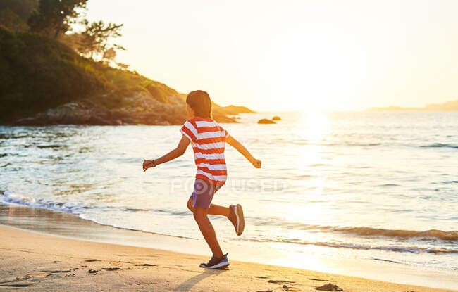 Side view full length of boy running on sandy shore washed by foamy sea — Stock Photo