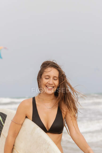 Joven deportista alegre con pelo volador y tabla de surf en el océano con espuma bajo el cielo nublado - foto de stock