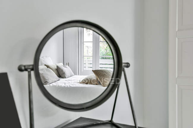 Круглое косметическое зеркало, расположенное на тумбочке и отражающее удобную кровать с подушками в светлом помещении — стоковое фото