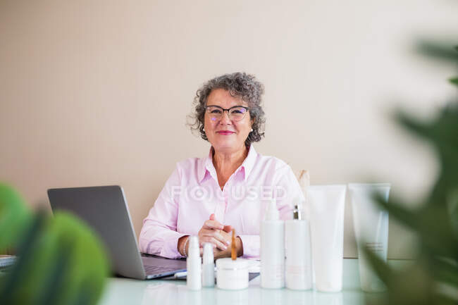 Улыбающаяся пожилая предпринимательница в очках смотрит в камеру на различные косметические средства и нетбук на светлом фоне — стоковое фото