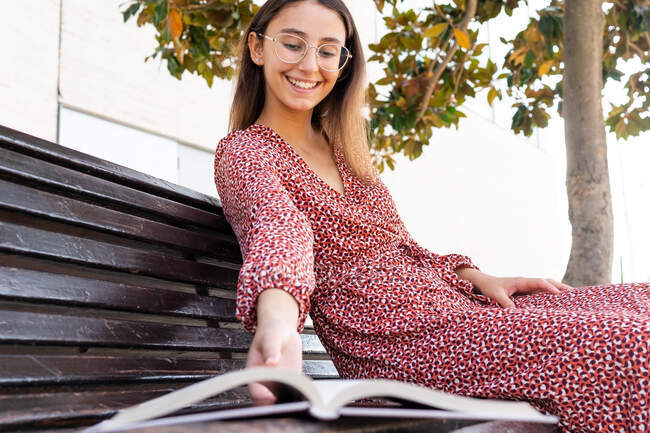 Dal basso di raccolto sorridente femminile in abito rosso seduto su una panchina di legno e prendendo aperto libro in strada durante il giorno — Foto stock