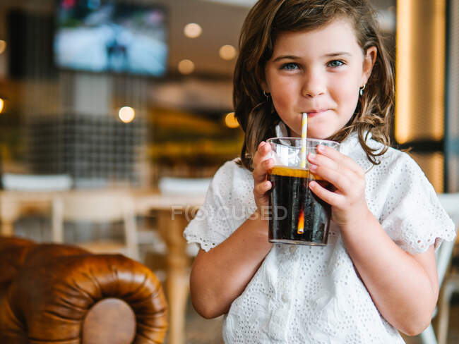 Cortar encantadora menina beber refrigerante carbonatado adoçado de vidro com palha e olhando para a câmera — Fotografia de Stock