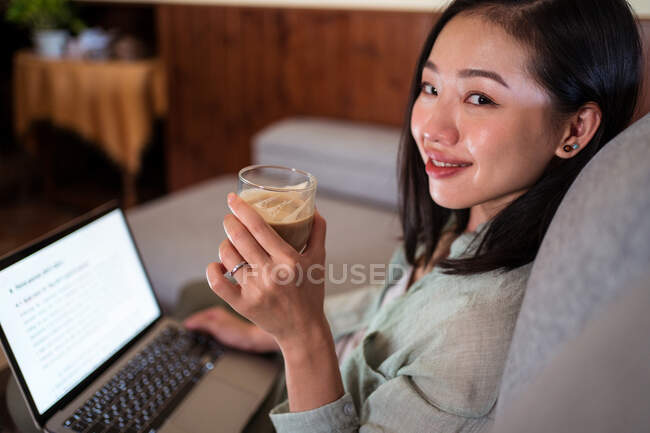 Side view of interested young ethnic female remote employee working on netbook on sofa drinking coffee at home looking at camera — Stock Photo