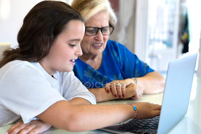 Вид збоку усміхненої онуки і бабусі, що сидить за столом і використовує ноутбук у світлій кімнаті в квартирі — стокове фото