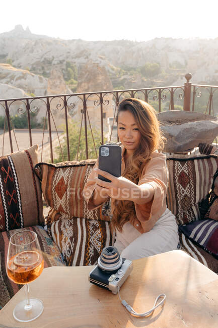 Alto ángulo de hembra étnica sentada en almohadas mientras toma autorretrato en el teléfono celular cerca de la mesa con cámara de fotos instantánea y cóctel en la terraza en Capadocia, Turquía - foto de stock