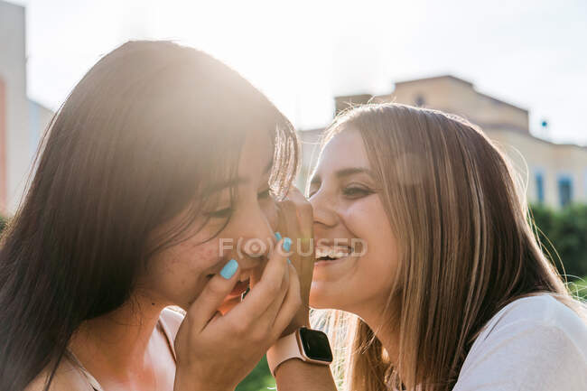 Cheerful teen covering mouth while whispering in ear of best female friend with closed eyes in sunlight — Stock Photo