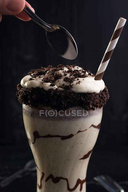 Crop anonymous person with spoon over glass of yummy milkshake decorated with crushed chocolate biscuits on black background — Stock Photo