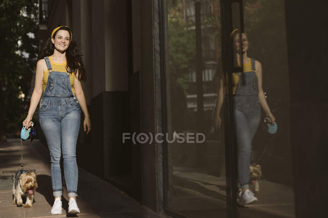 Full body of positive female owner enjoying walk with small Yorkshire Terrier dog with tongue out near reflecting wall — Stock Photo