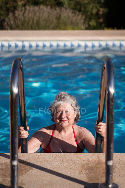 Позитивна старша жінка в купальнику спускається в басейн і тримає нержавіючі поручні під час відпочинку в сонячний день — стокове фото