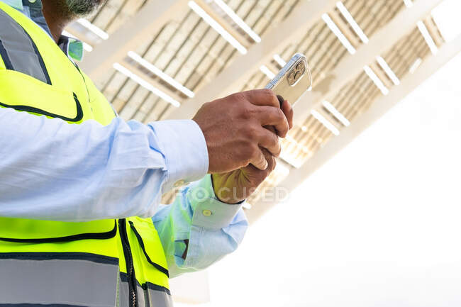 Cropped unrecognizable foreman in waistcoat browsing on smartphone while standing near solar power station — Stock Photo