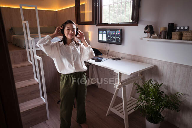 Young ethnic smiling female trader putting on wireless headphones looking at camera before analyzing cryptocurrency graphics on computer monitor in house — Stock Photo
