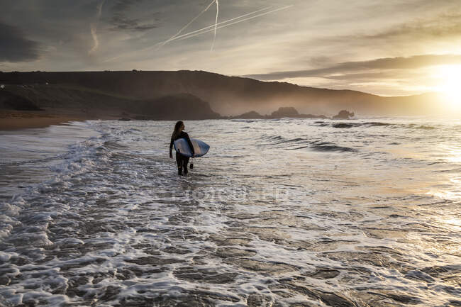 Visão traseira da jovem com prancha de surf entrando no mar durante o pôr do sol na praia em Astúrias, Espanha — Fotografia de Stock