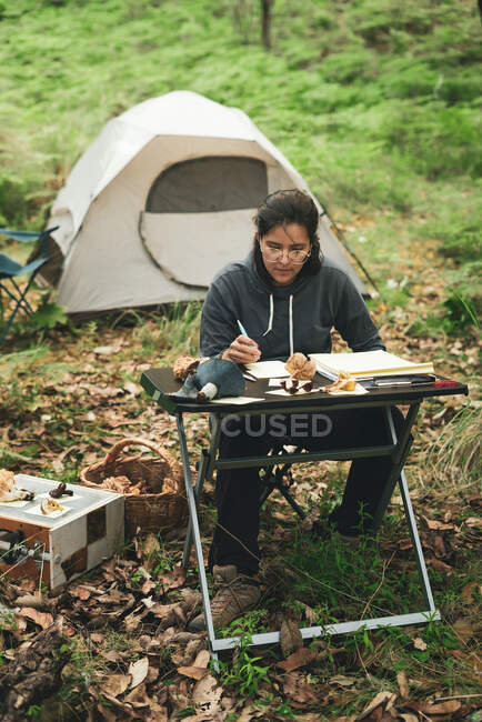 Thoughtful female in casual clothes and eyeglasses sitting at table and taking notes in notebook while researching mushrooms in green park against tent in daytime — Stock Photo