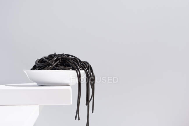 Minimalistisches Atelier mit Spaghetti mit schwarzer Tintenfischtinte, die aus einer vollen Keramikschale auf weißem Tisch herausfallen — Stockfoto