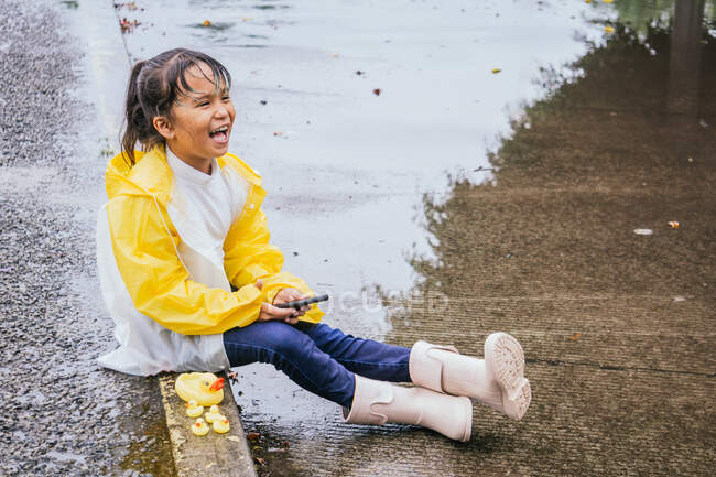 Criança étnica despreocupada em capa de chuva com celular descansando na passarela contra patos de brinquedo enquanto ri e olha para a frente em dia chuvoso — Fotografia de Stock
