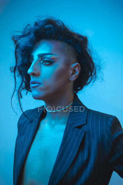 Young transsexual male model with makeup in stylish jacket looking away on blue background — Stock Photo