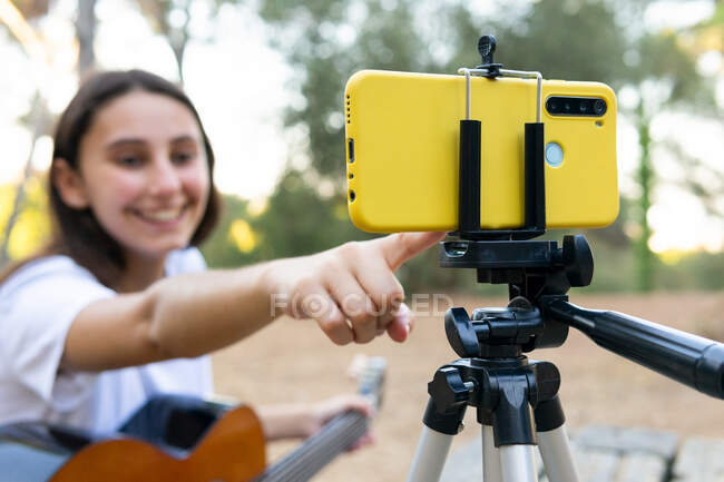 Усміхнена жінка підліток гітарист записує відео на сучасному мобільному телефоні на тринозі в парку на розмитому фоні — стокове фото