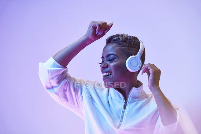 Mujer étnica enérgica en auriculares inalámbricos y ropa de moda bailando hip hop con la boca abierta y los ojos cerrados - foto de stock