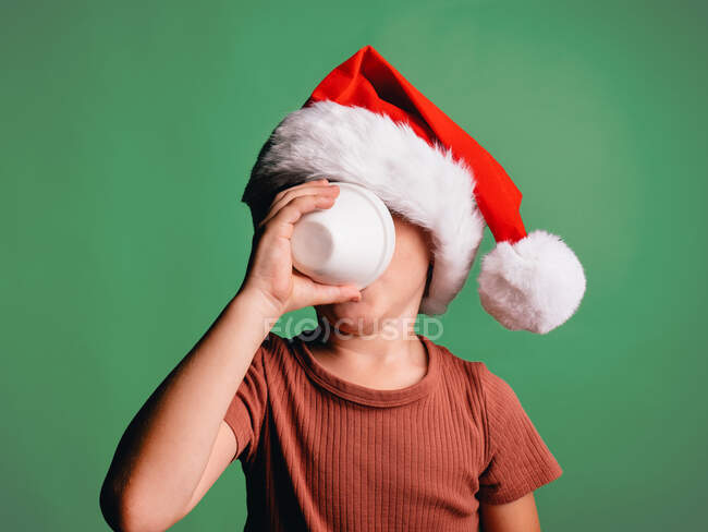 Kleiner Junge mit rotem Weihnachtsmann zur Feier des neuen Jahres trinkt aus Tasse, während er auf grünem Hintergrund steht — Stockfoto