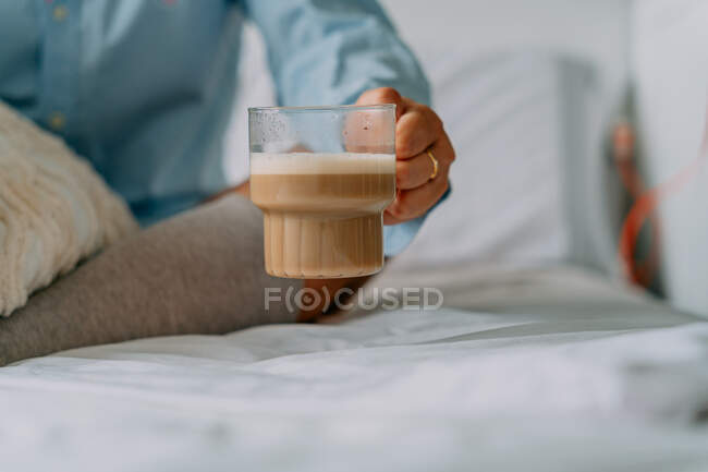 Coltivazione femminile irriconoscibile in anello con tazza di delizioso caffè con schiuma di latte in camera da letto — Foto stock