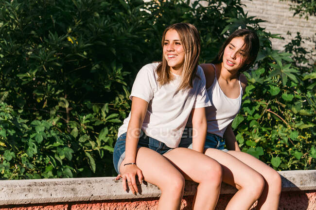 Adolescent joyeux avec contemplative meilleure amie regardant loin tout en étant assis sur un banc urbain au soleil — Photo de stock