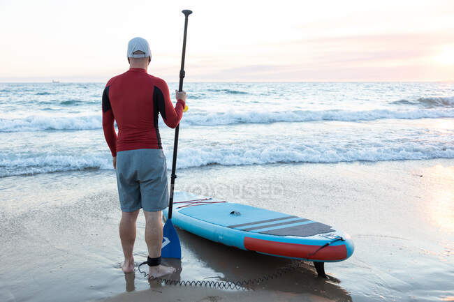 Back view of unrecognizable male surfer in wetsuit and hat standing with paddle and SUP board while preparing to surf on seashore — Stock Photo