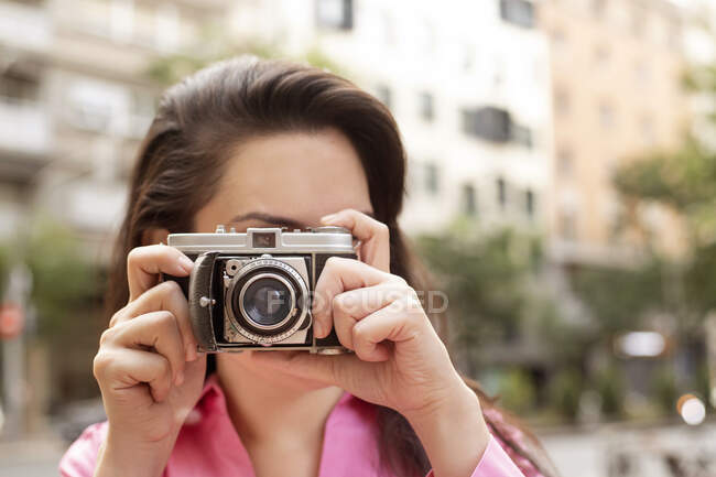 Young female with long brown hair taking picture on old fashioned photo camera on street in city — Stock Photo