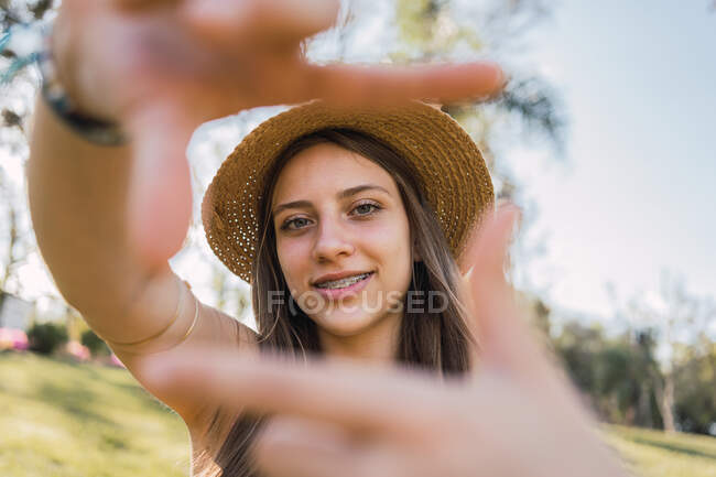 Smiling female teenager with braces demonstrating photography gesture while looking at camera in daytime on blurred background — Stock Photo