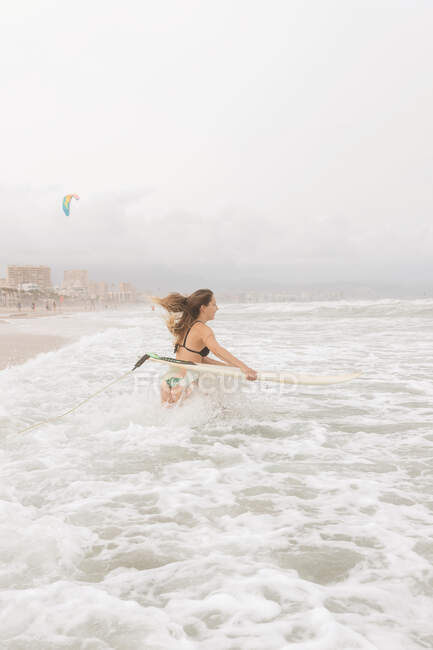 Vista lateral de la joven deportista con pelo volador y tabla de surf en el océano con espuma bajo el cielo nublado - foto de stock