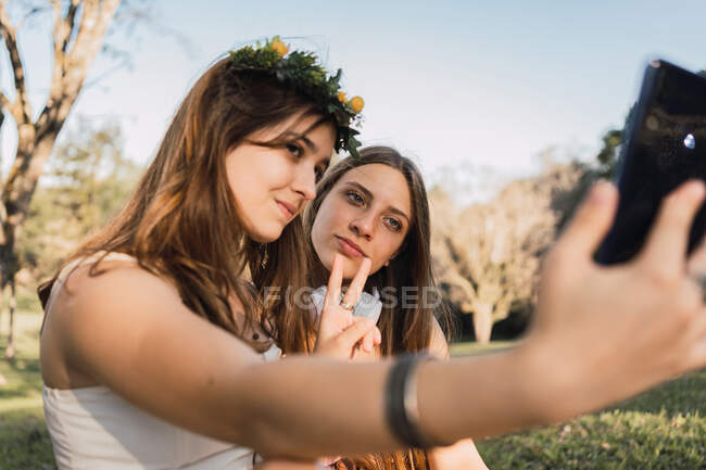 Подростки демонстрируют победный жест, делая автопортрет на мобильном телефоне в солнечном парке на размытом фоне — стоковое фото